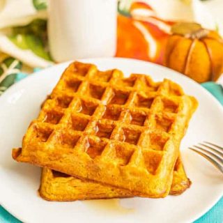 Two large pumpkin waffles stacked on a white plate with maple syrup on top and a fork to the side
