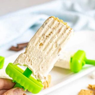 A hand holding a homemade s'mores popsicle with a green stick base and graham cracker crumbs on the top