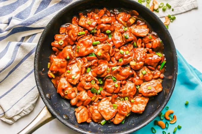 Cashew chicken in a large skillet with green onions on top and green onions and cashews sprinkled on the counter nearby