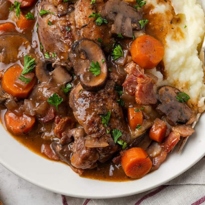 Close up of coq au vin served over mashed potatoes on a white plate with a fork resting on the side.